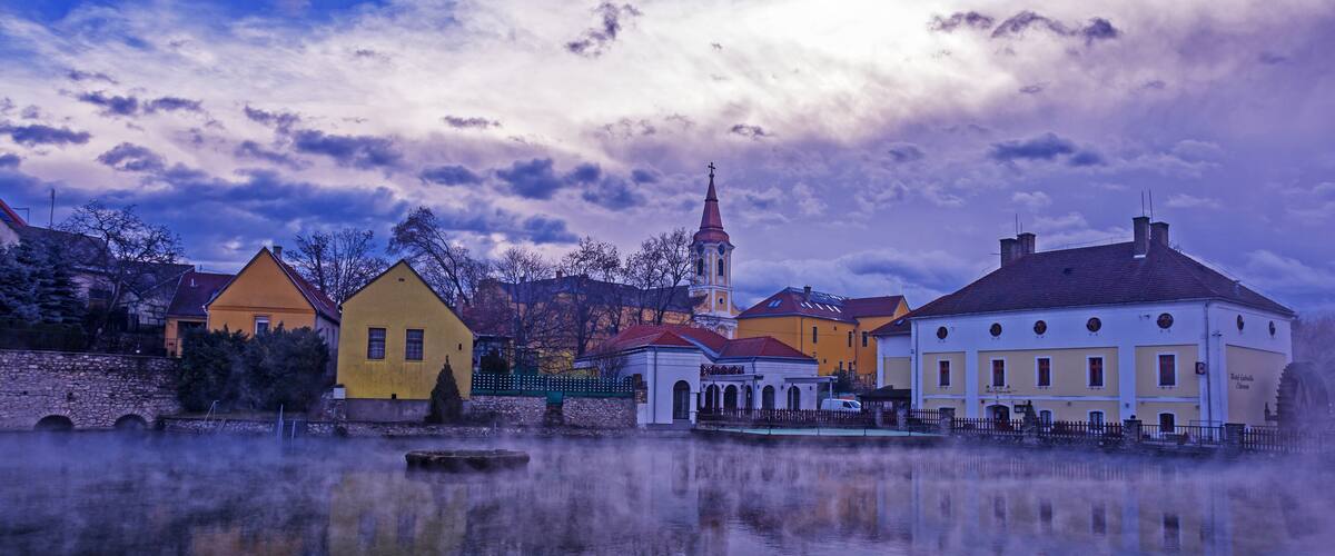 A calm lake reflecting buildings, a church with a tall tower, and a stone wall.