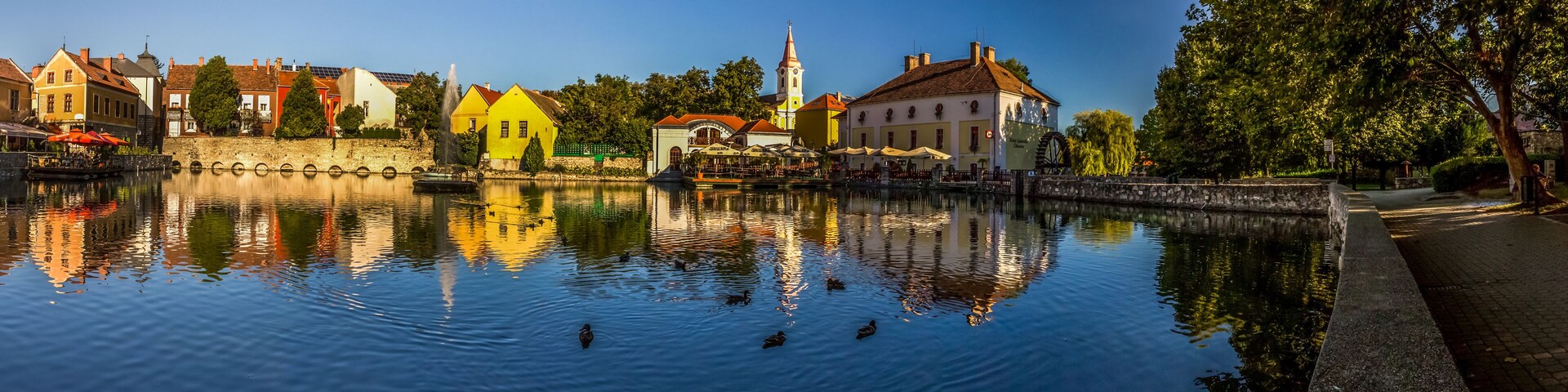 Un río tranquilo que refleja edificios de colores, una fuente y patos nadando.