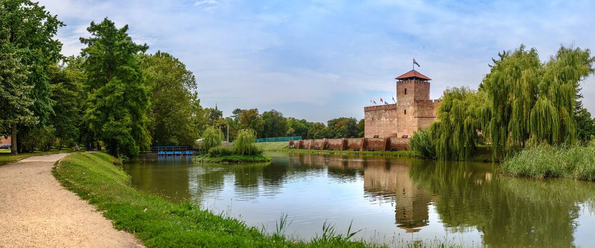 A historic castle by a calm river, surrounded by lush greenery and trees.