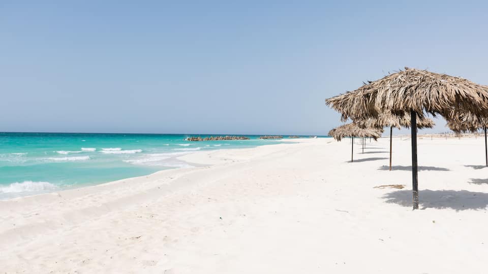 A beach with white sand, turquoise water, and thatched umbrellas.