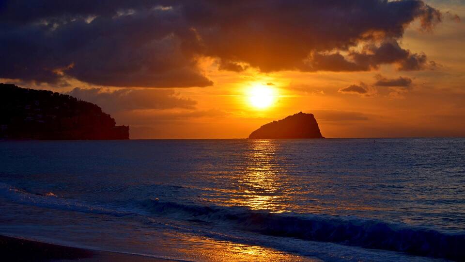 A sunset over the ocean with a silhouette of a landmass and waves on the shore.
