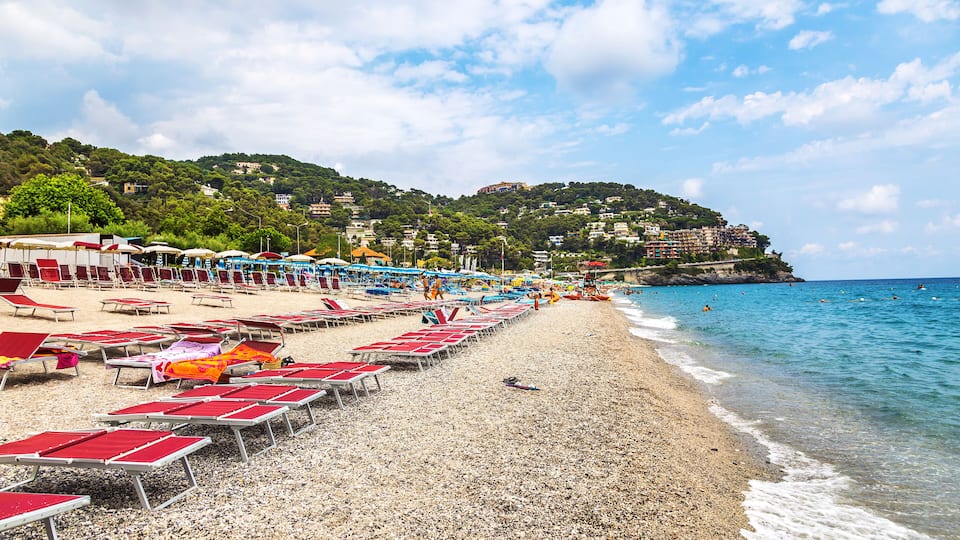 A pebble beach with red and white sun loungers, a clear blue sea, and buildings on a hillside in the background.