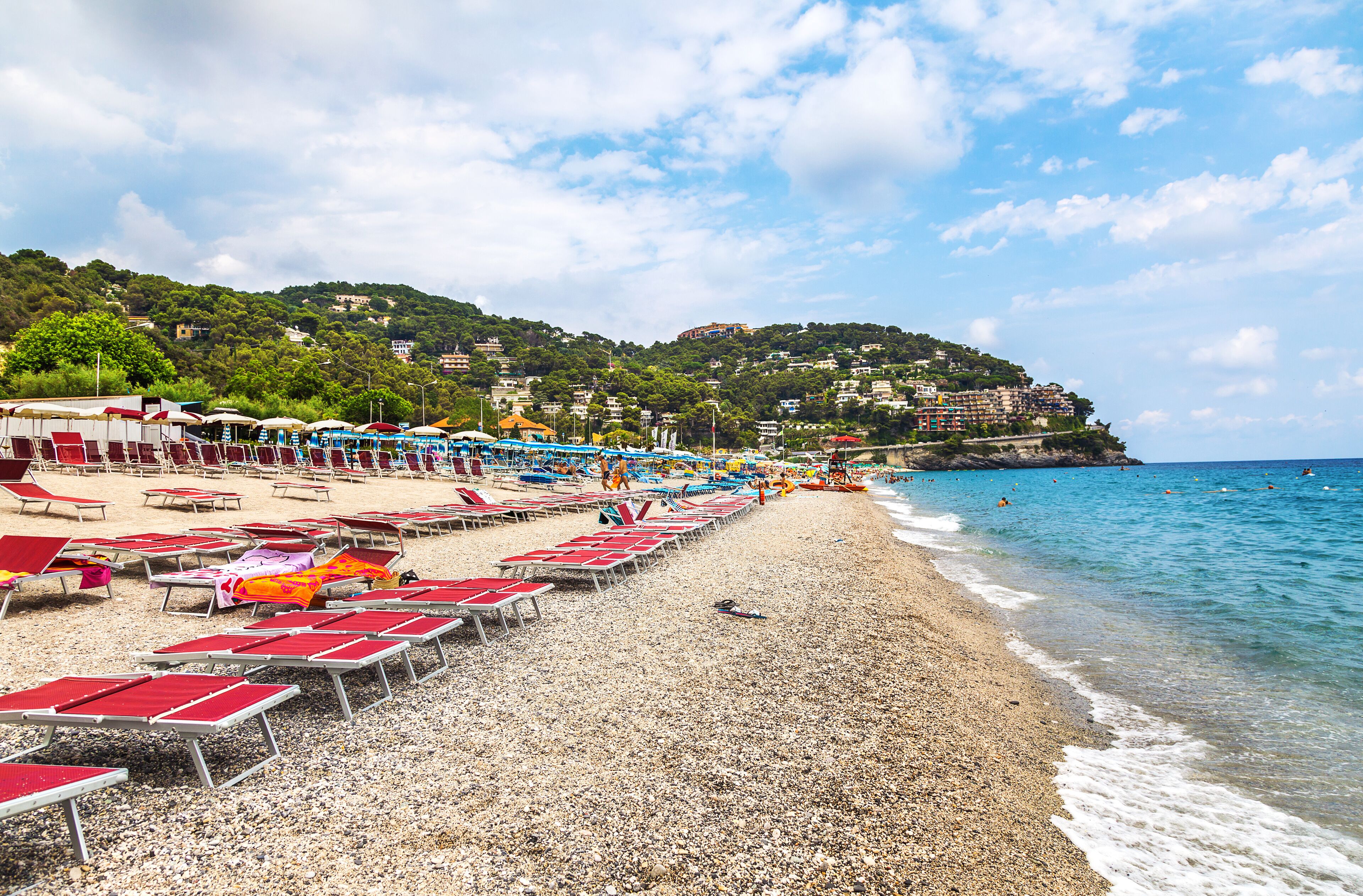 A pebble beach with red and white sun loungers, a clear blue sea, and buildings on a hillside in the background.