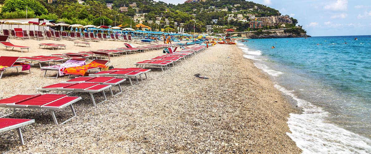A pebble beach with red and white sun loungers, a clear blue sea, and buildings on a hillside in the background.