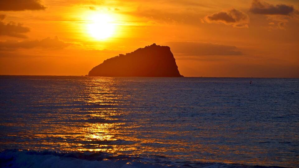 A sunset over the ocean with a prominent rock formation silhouetted against the sky.
