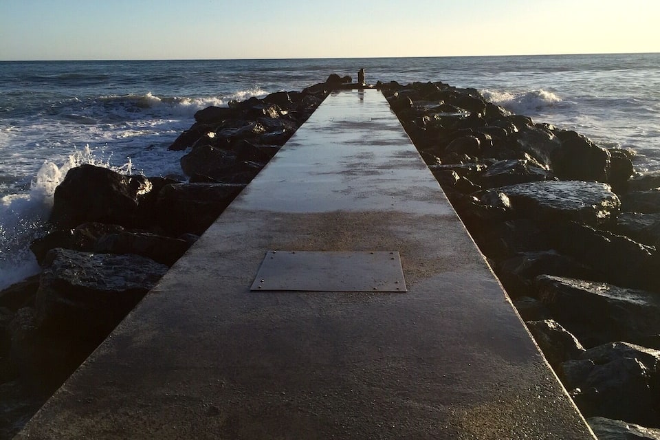 A concrete pier extending into the ocean with waves crashing against rocks on both sides.