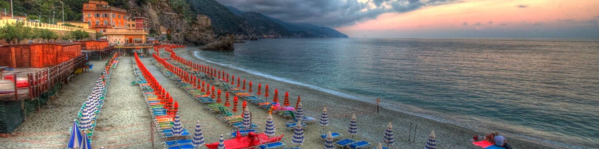 A beach with rows of blue and red umbrellas, a calm sea, and a mountainous backdrop.
