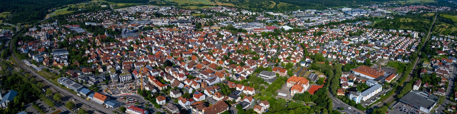 An aerial view of a densely populated town with red-roofed houses, surrounded by green hills and a clear sky.