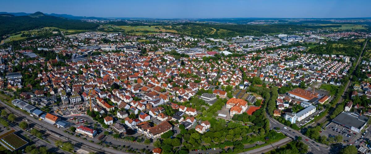 An aerial view of a densely populated town with red-roofed houses, surrounded by green hills and a clear sky.