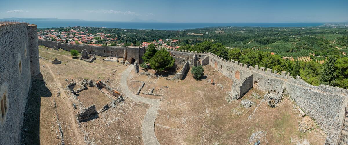 An ancient stone fortress with ruins, a clear view of the surrounding landscape, and a distant view of a coastal town.