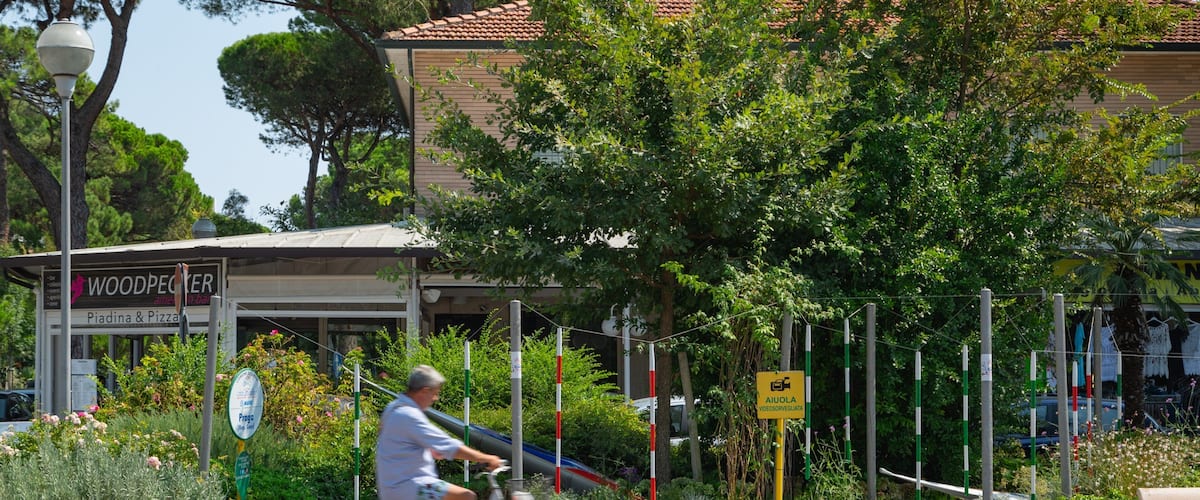 A man riding a bicycle past a restaurant named "WOODPECKER" and a sign for "AIOULA VIDEOSORVEGIATA."