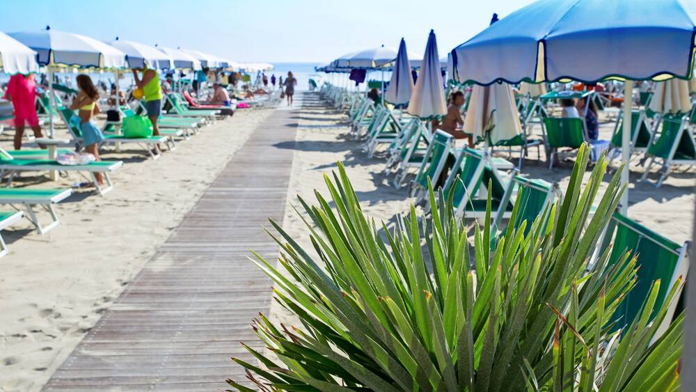 Wooden walkway on the sand to the sea. Umbrellas and couches on blue sky and sea background on the beach of Milano Marittima, Adriatic coast, Italy. Popular Tourist Resort at Adriatic Sea