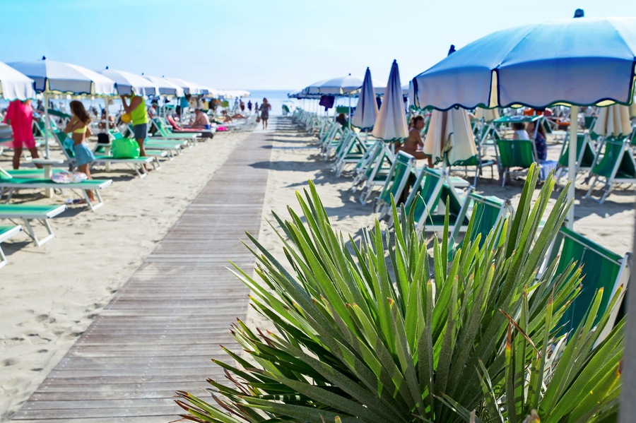 Wooden walkway on the sand to the sea. Umbrellas and couches on blue sky and sea background on the beach of Milano Marittima, Adriatic coast, Italy. Popular Tourist Resort at Adriatic Sea