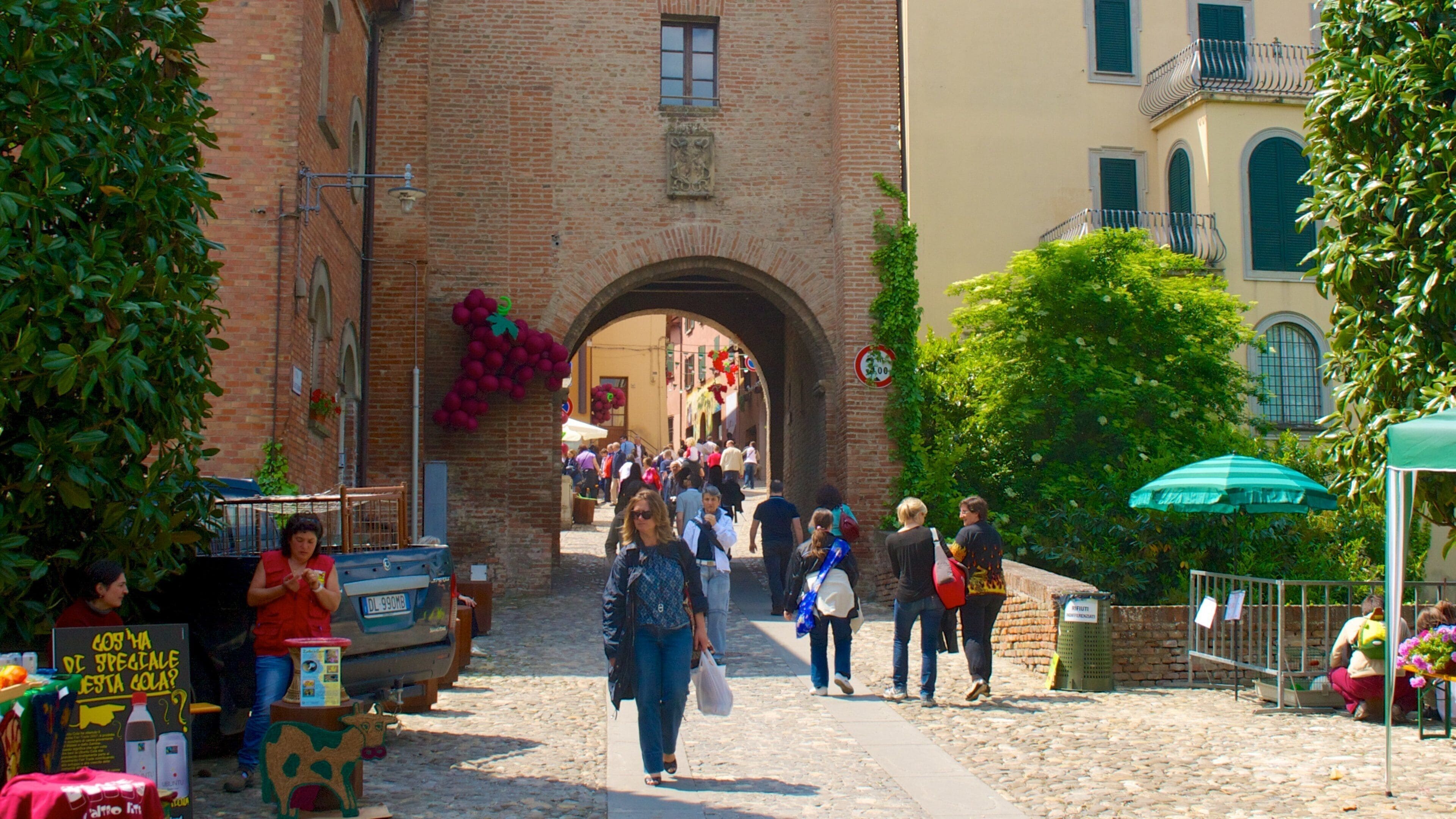 A cobblestone street with a brick archway, people walking, and a market stall with a sign.