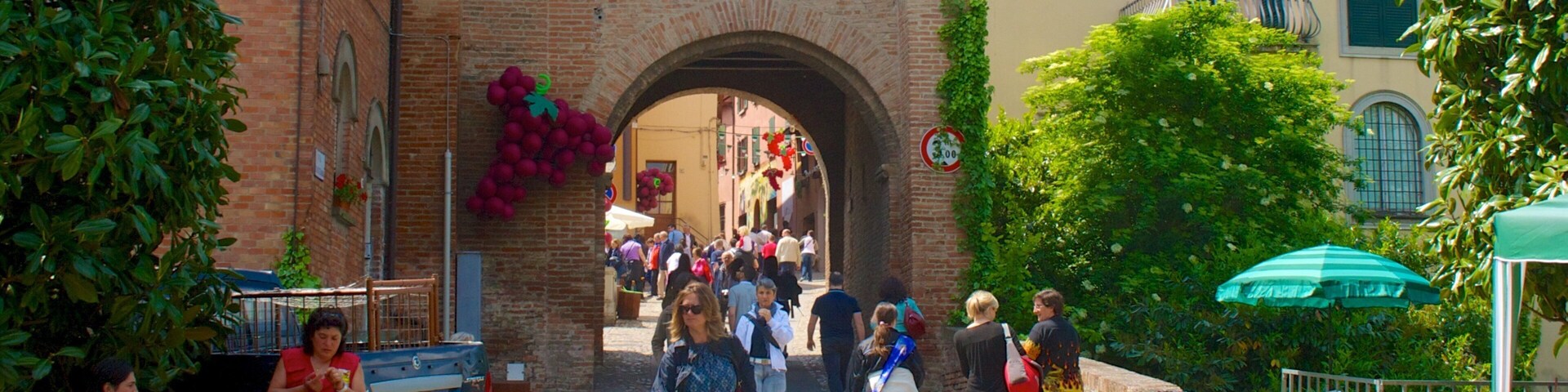 Una calle empedrada con un arco de ladrillo, gente caminando y un puesto de mercado con un letrero.