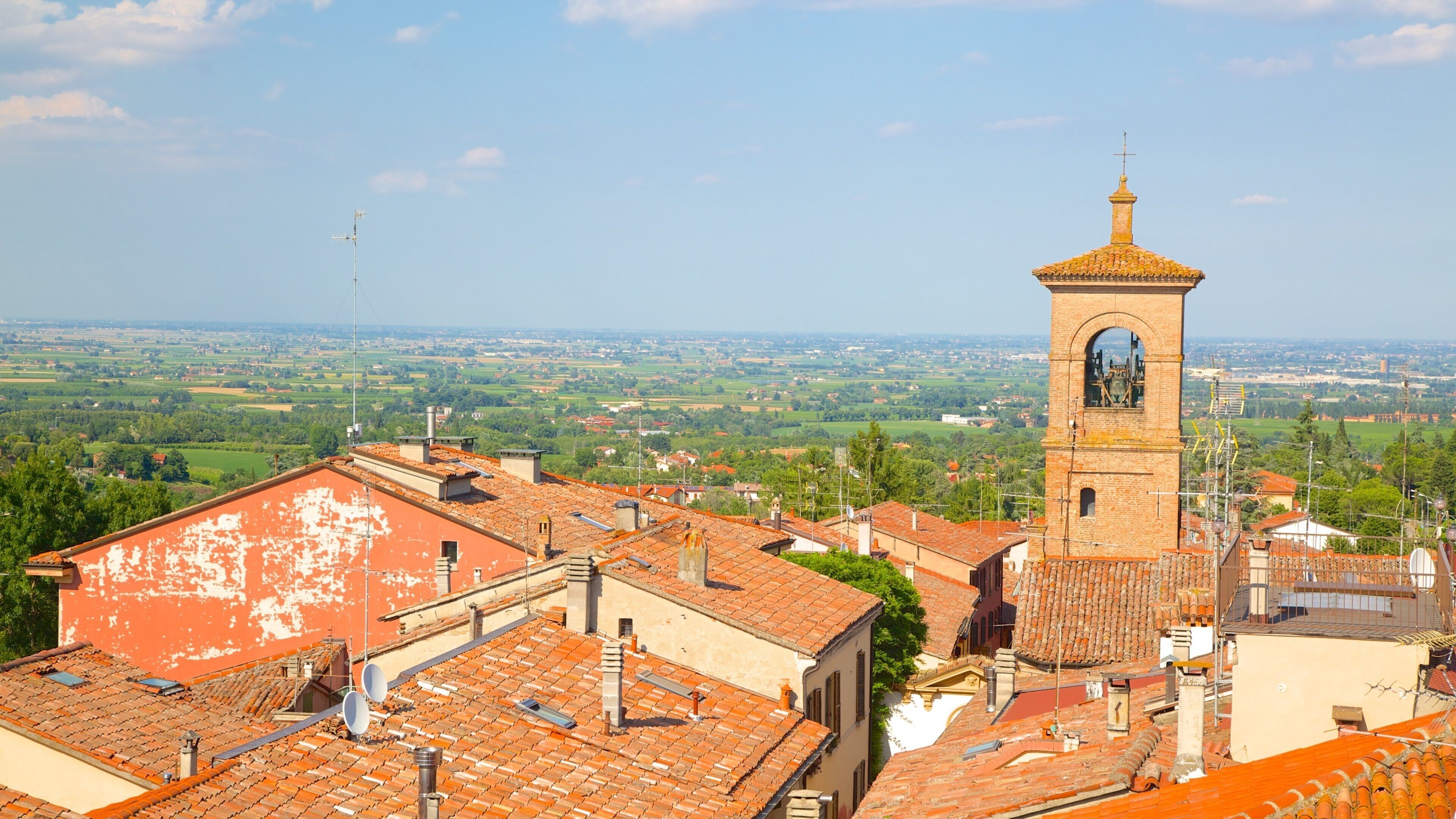 A town with a prominent tower, terracotta-roofed buildings, and a vast green landscape in the background.