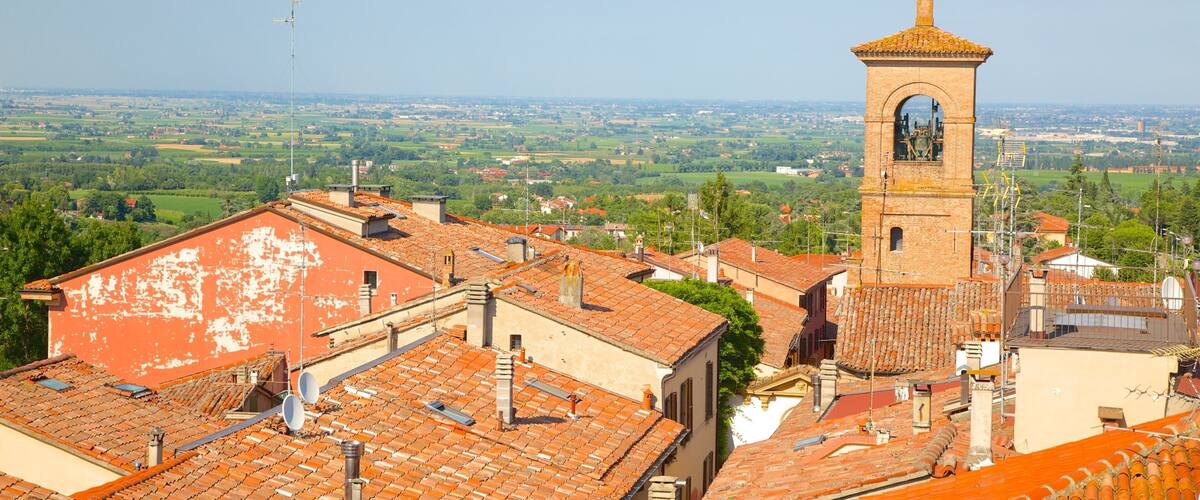 A town with a prominent tower, terracotta-roofed buildings, and a vast green landscape in the background.