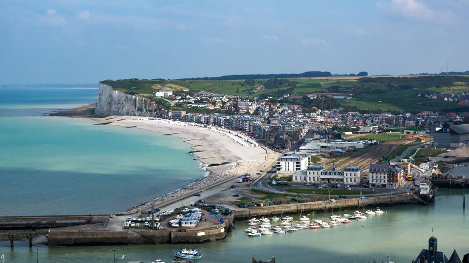 A coastal town with a long sandy beach, a harbor with boats, and a cliff overlooking the sea.