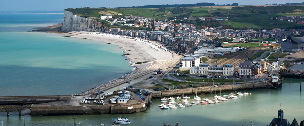 A coastal town with a long sandy beach, a harbor with boats, and a cliff overlooking the sea.