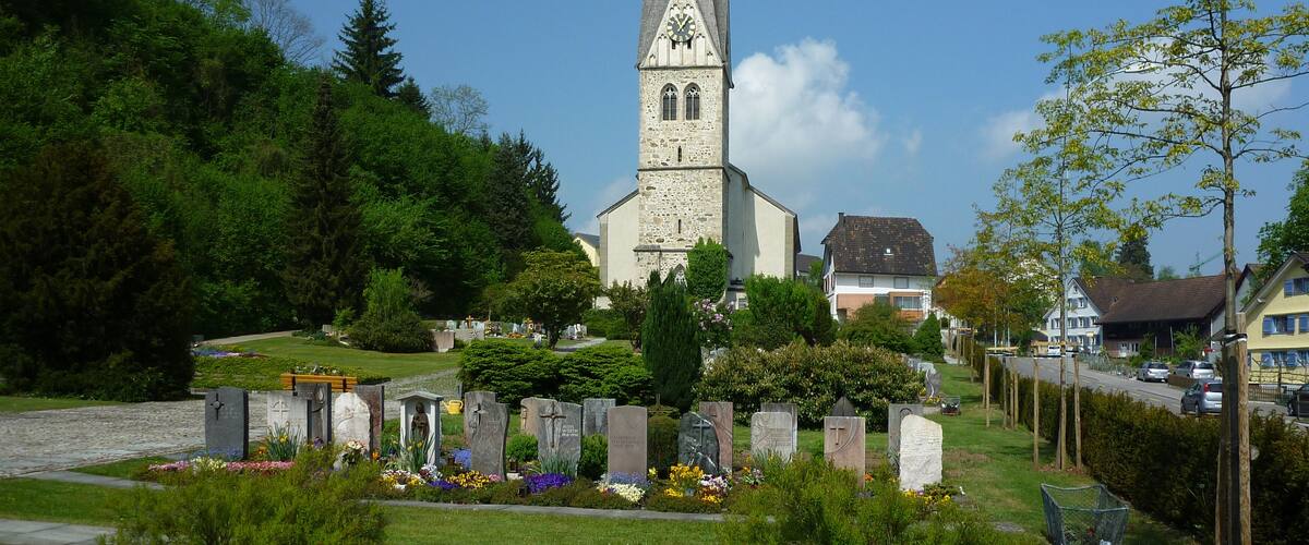 A church with a tall spire, surrounded by a cemetery with headstones and flowers.