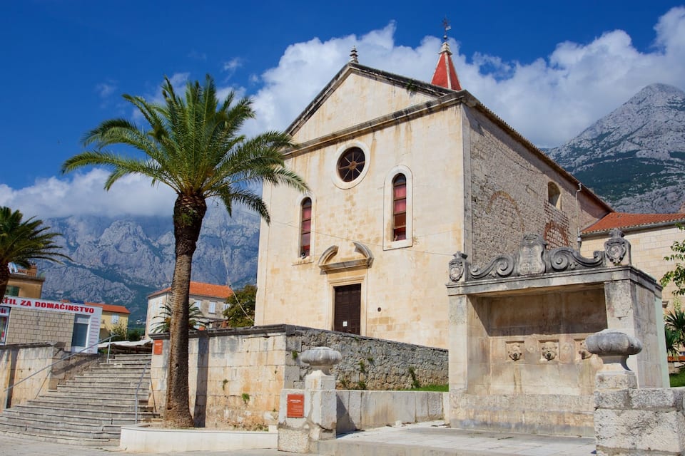 A historic church with a red roof, palm trees, and mountains in the background.