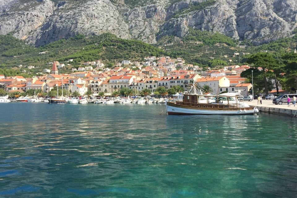 A coastal town with clear turquoise waters, a wooden boat docked, and rocky mountains in the background.
