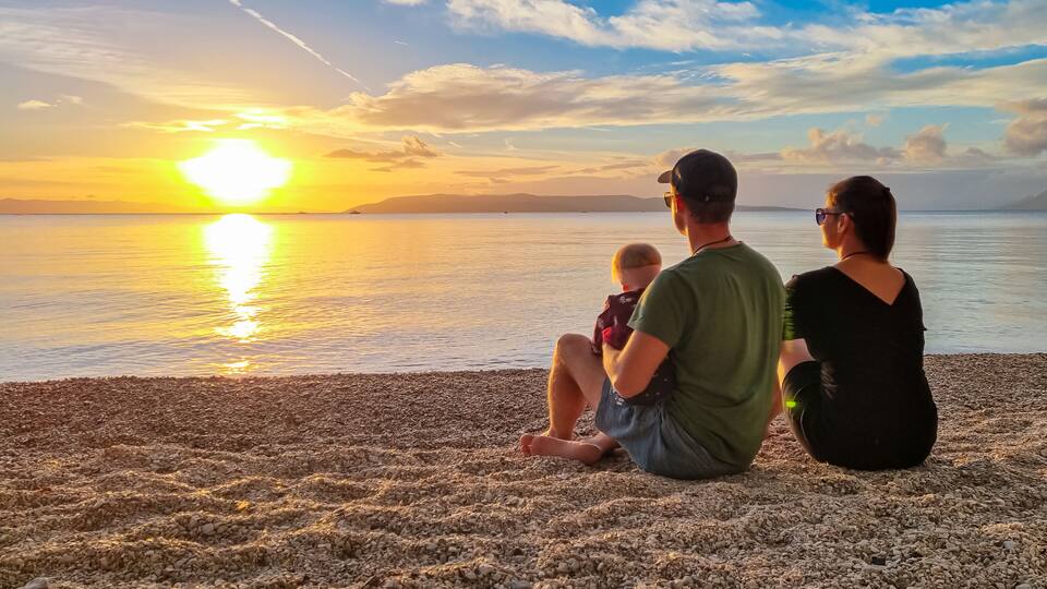 A family sitting on a pebbled beach at sunset with a calm sea and distant mountains.