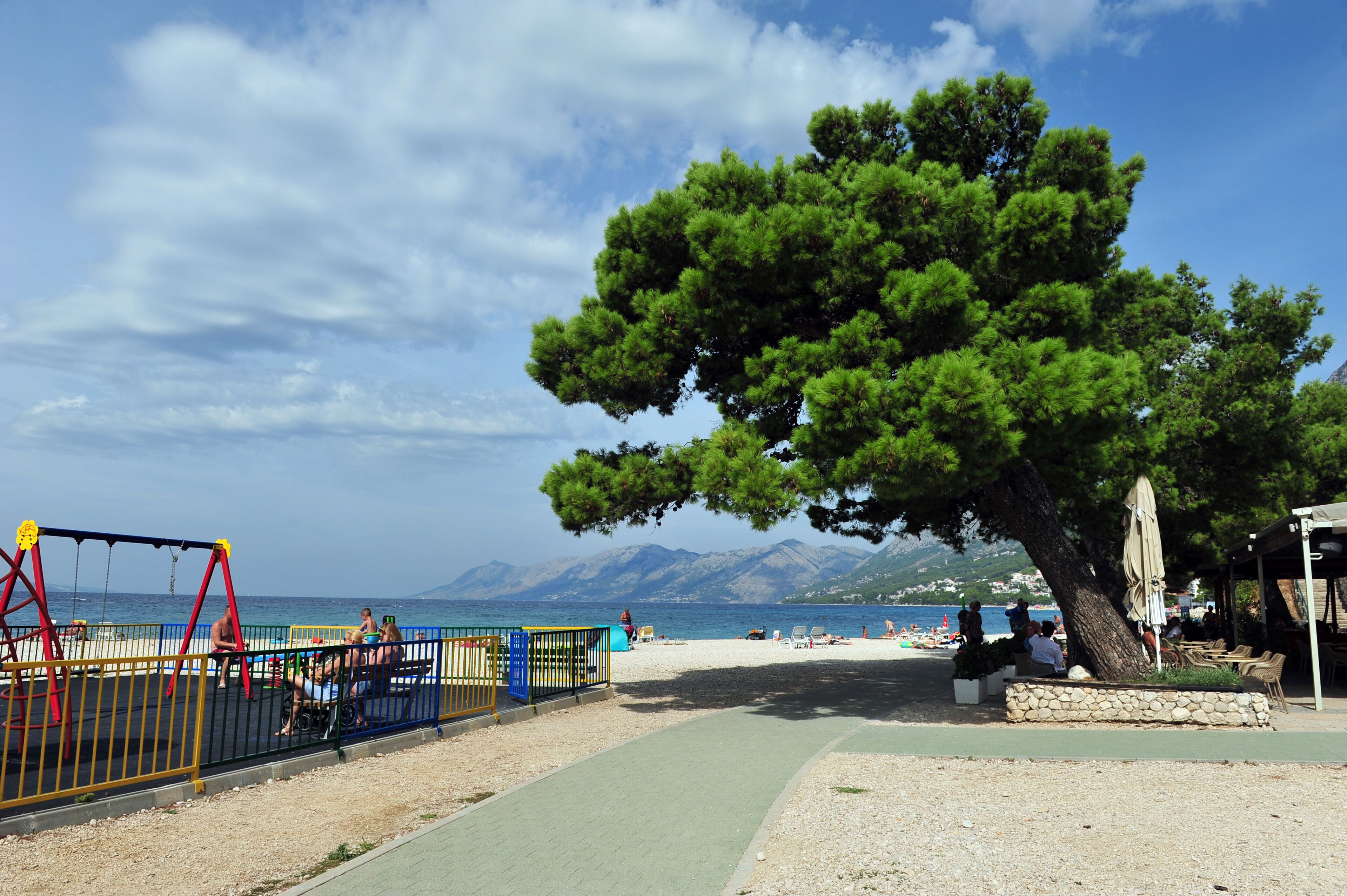 Una playa con un parque infantil, un árbol grande y una vista despejada del mar y las montañas al fondo.