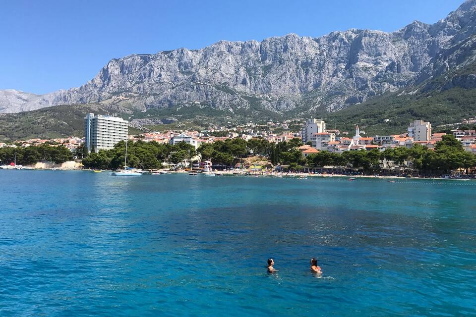 A coastal town with a mountain backdrop, clear blue waters, and a few people swimming.