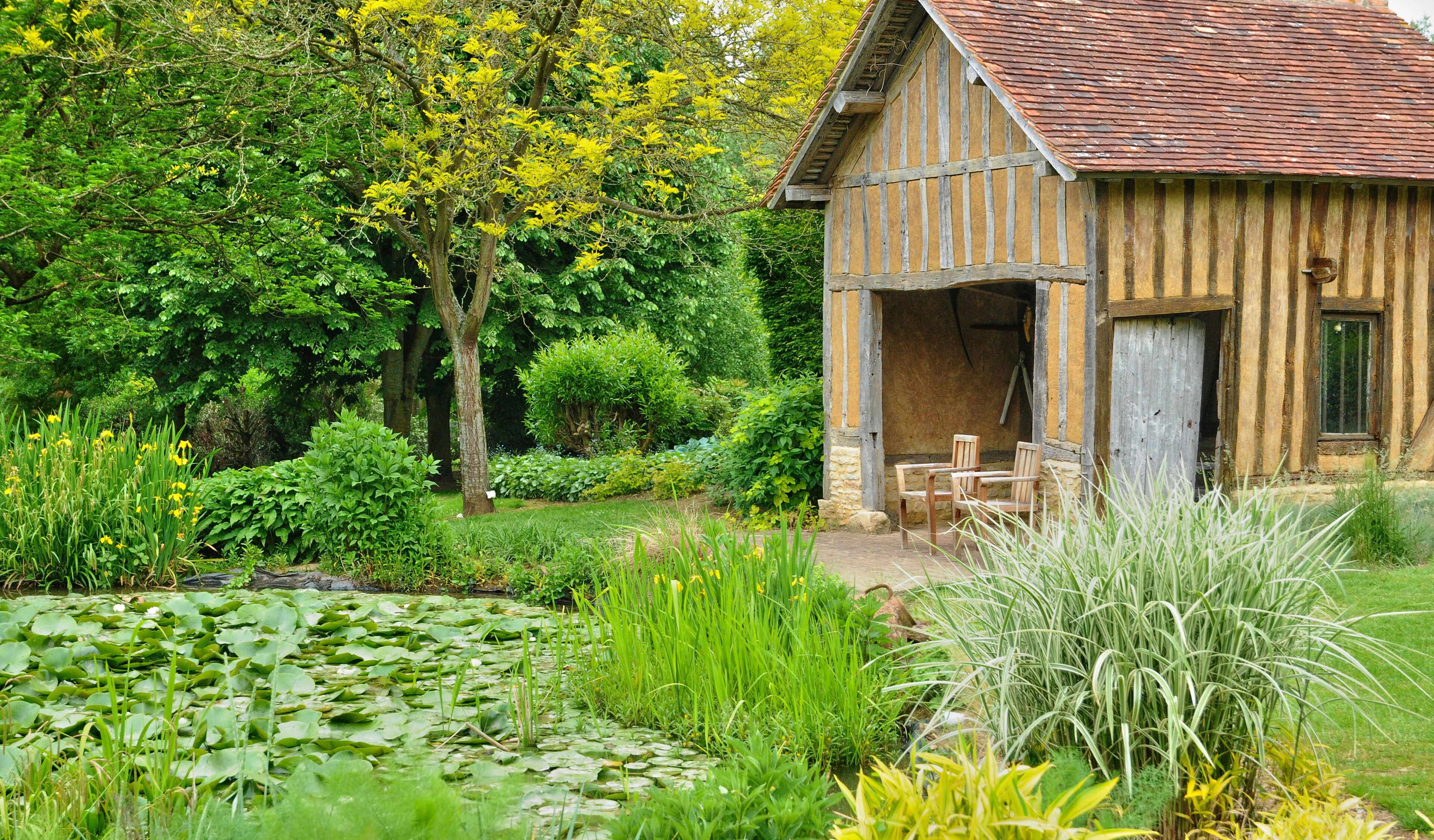 A rustic wooden house with a tiled roof, surrounded by lush greenery and a pond with lily pads.