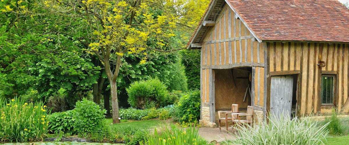 A rustic wooden house with a tiled roof, surrounded by lush greenery and a pond with lily pads.