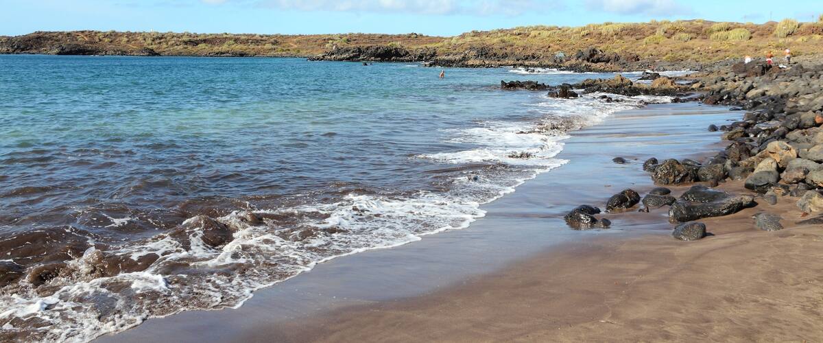 A beach with waves, rocky shore, and distant hills.