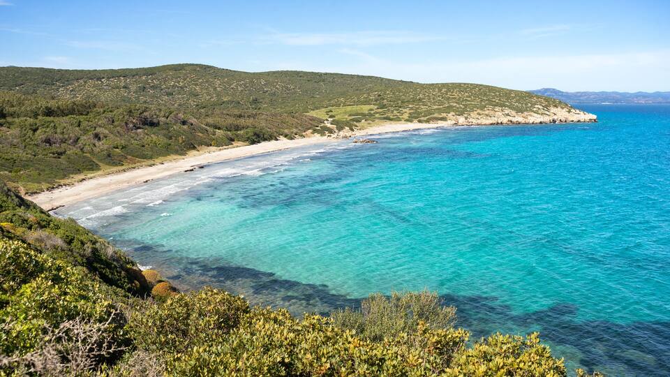 A coastal landscape with clear turquoise waters, a sandy beach, and green hills in the background.