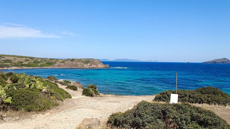 A coastal landscape with a clear blue sea, rocky cliffs, and sparse vegetation.