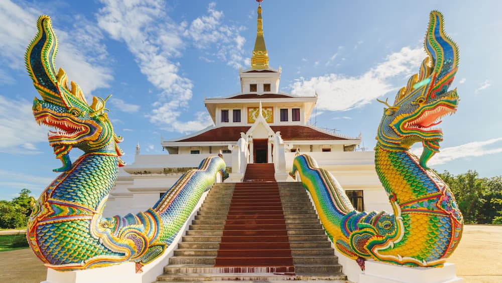 A pair of colorful Naga statues stand majestically atop the grand staircase of Wat Pha Tak Suea Temple in Sangkhom District, Nong Khai Province, Thailand, against a clear blue sky.