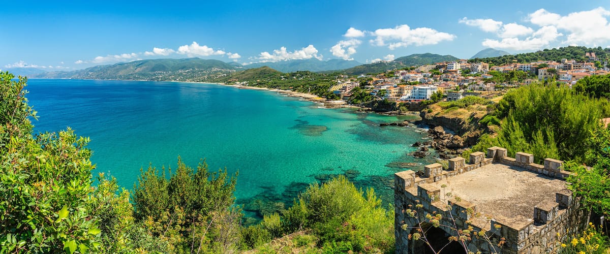 A coastal town with turquoise waters, a historic stone tower, and lush greenery.