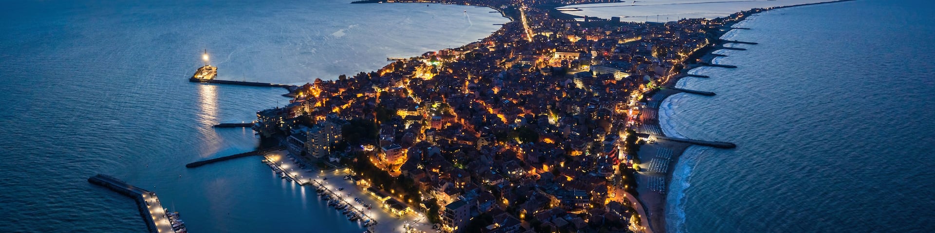 An aerial view of a coastal city at dusk with illuminated buildings and a prominent peninsula extending into the sea.