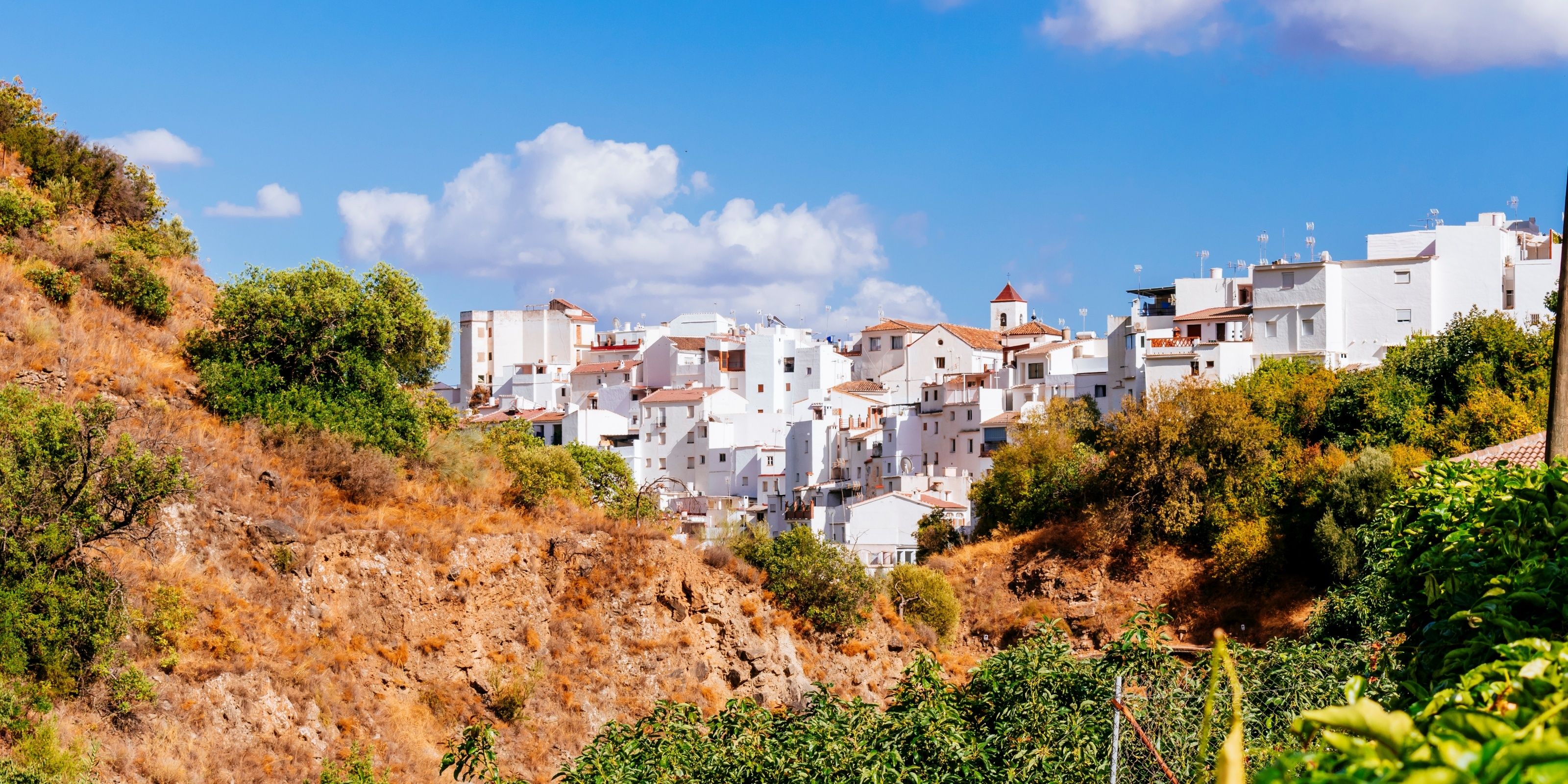 A hillside town with white buildings and red-tiled roofs, surrounded by dry vegetation and a clear blue sky.
