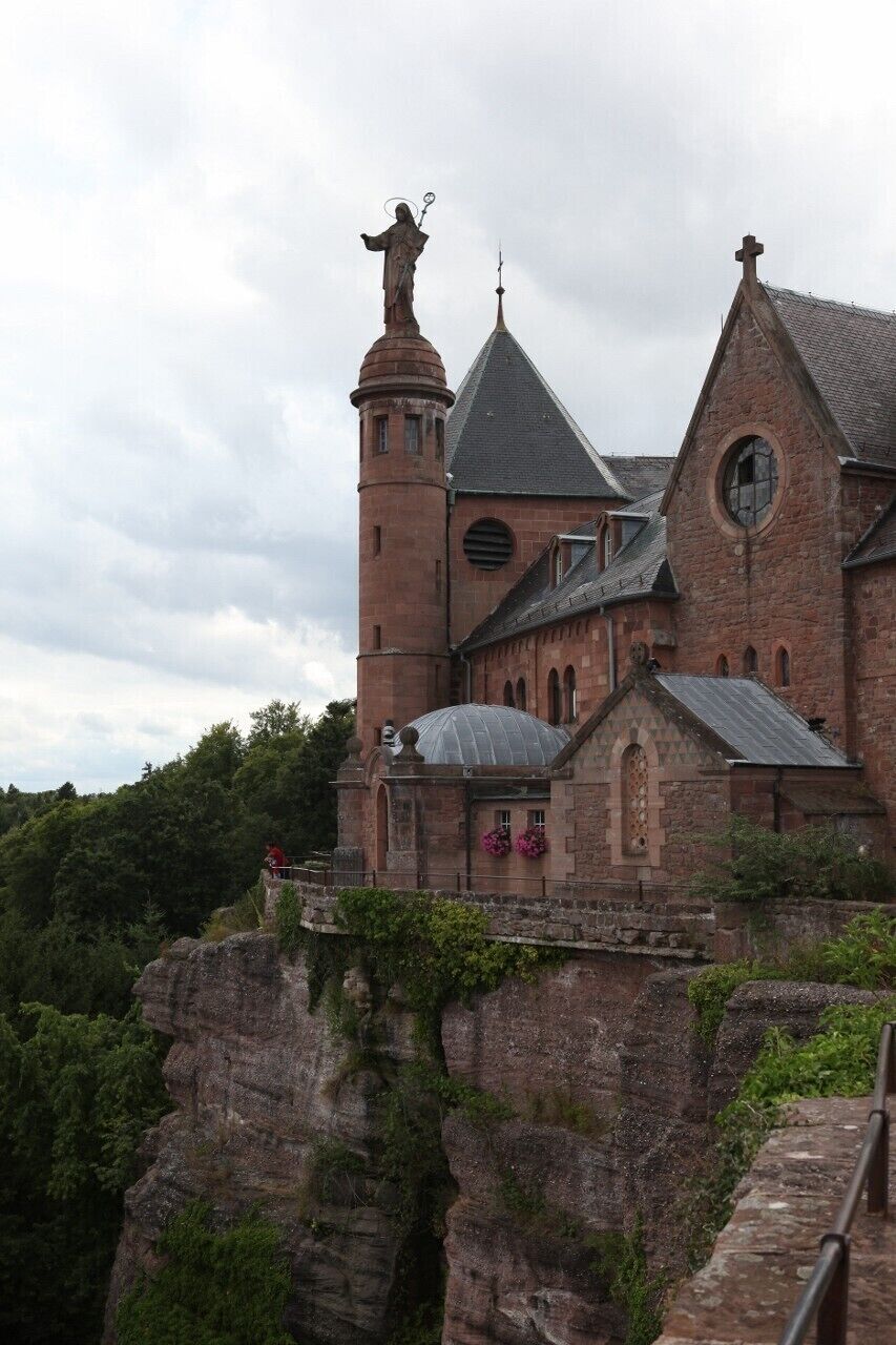 A historic church with a prominent bell tower and a statue atop it, situated on a cliff with lush greenery around.