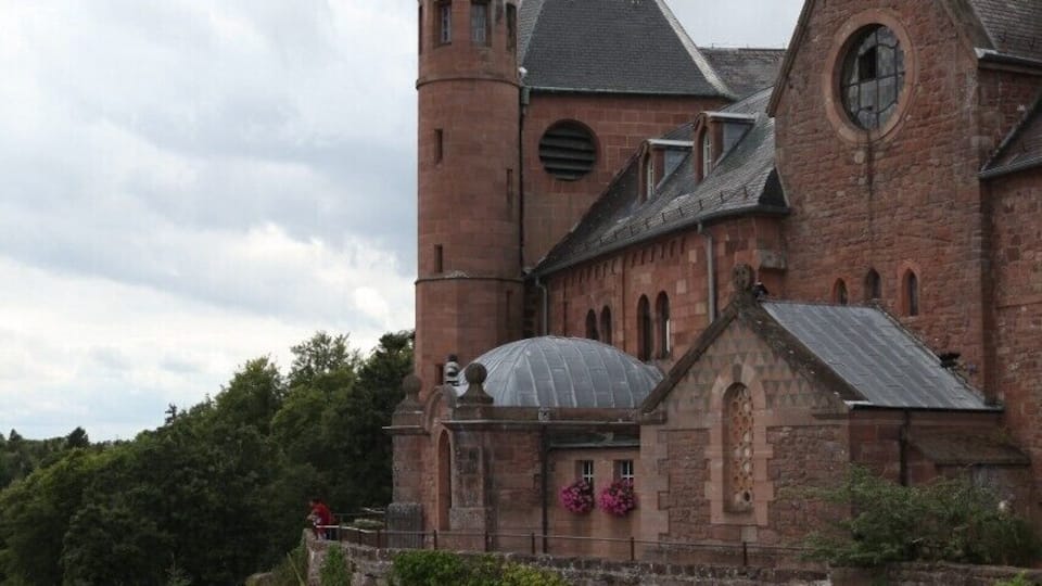 A historic church with a prominent bell tower and a statue atop it, situated on a cliff with lush greenery around.