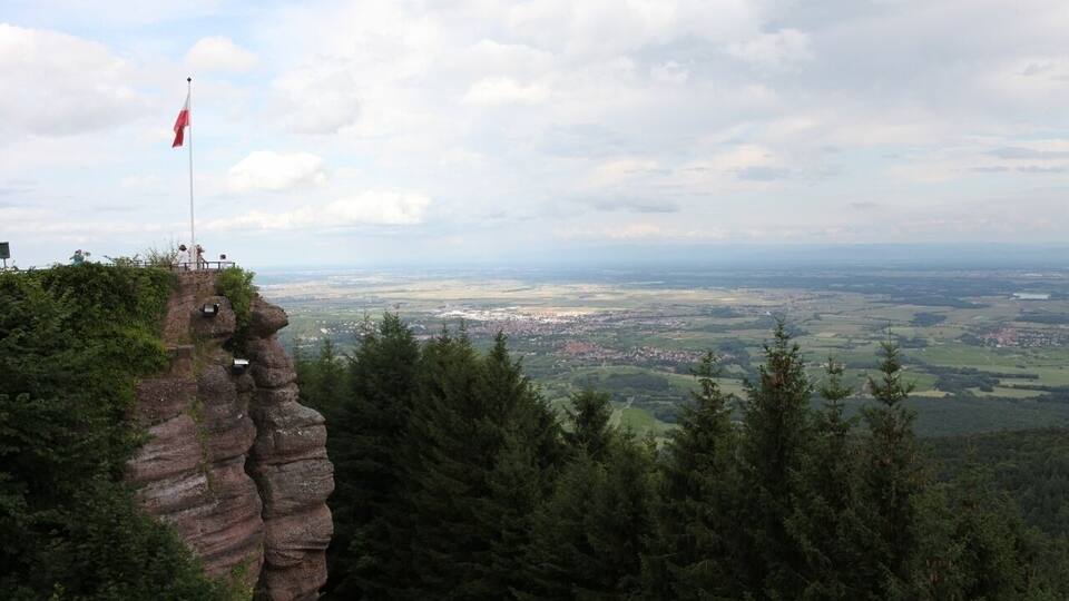 A flag on a rocky cliff overlooking a valley with a distant cityscape.