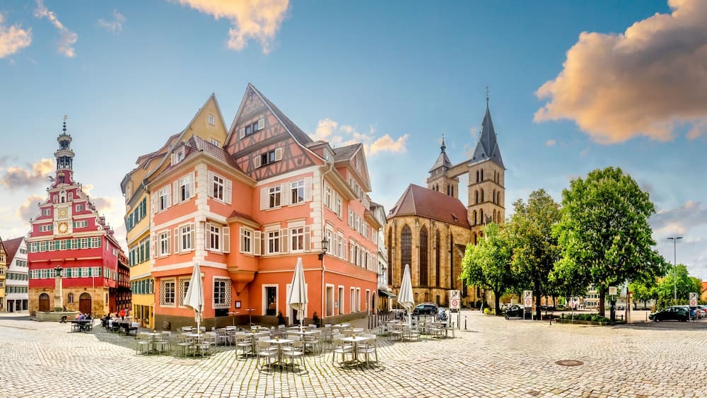 Une place pavée bordée de bâtiments aux façades à colombages aux couleurs vives, une église dotée d’un clocher imposant et un ciel dégagé.