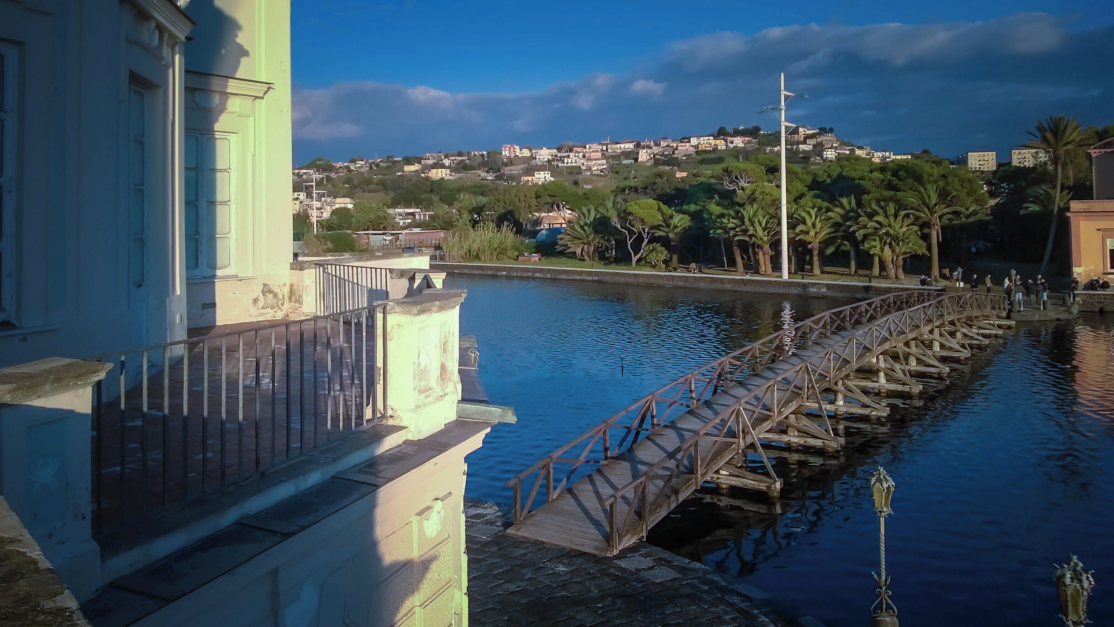 A wooden bridge over a calm river, with buildings and a hill in the background.