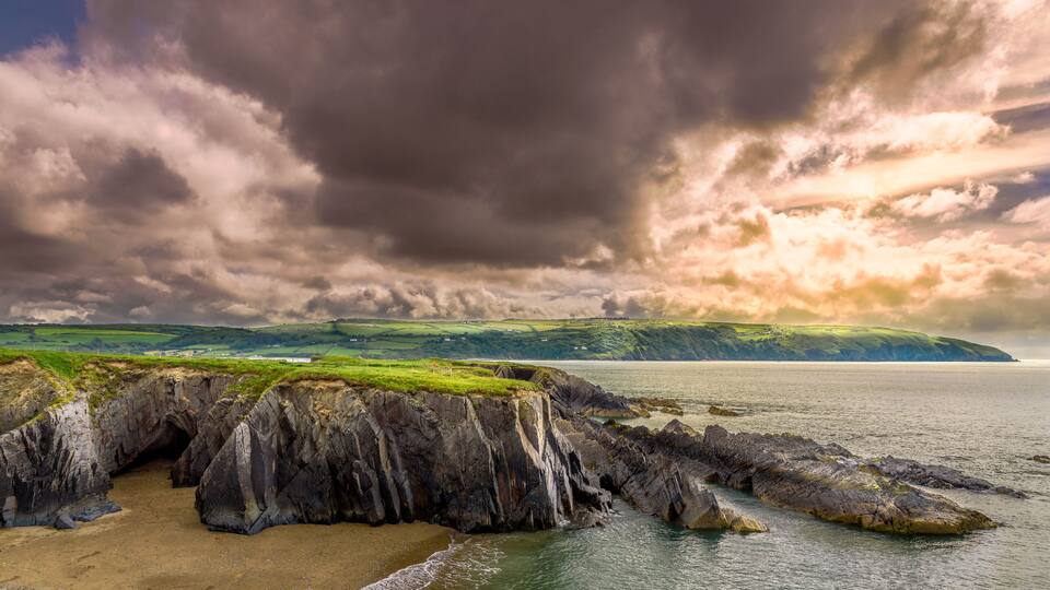 A coastal landscape with cliffs, a sandy beach, and a distant view of green hills under a cloudy sky.
