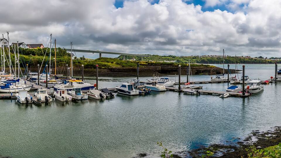 A marina with various boats docked, a bridge in the background, and a green hillside.