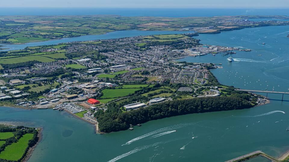 An aerial view of a coastal town with a marina, a bridge, and surrounding green fields.