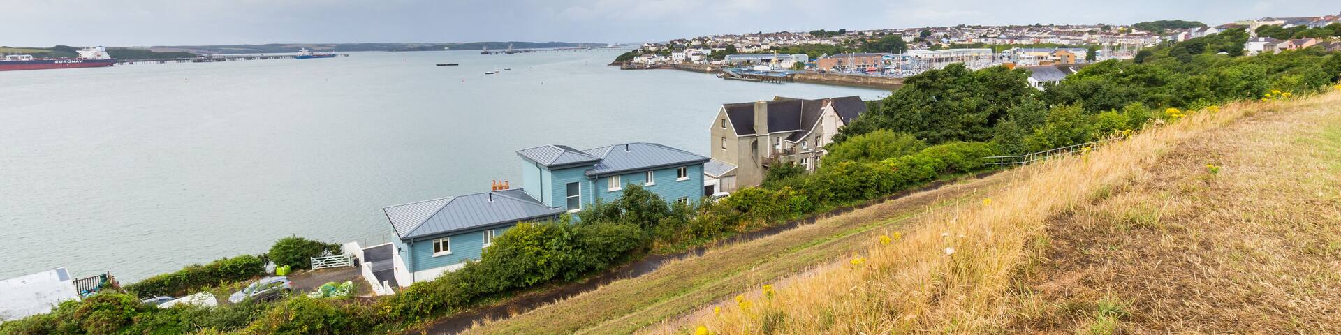 A coastal town with houses near a large body of water, a pier, and a bridge in the distance.