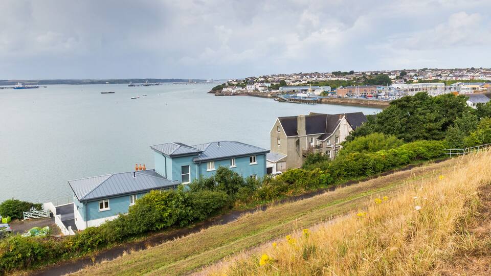 A coastal town with houses near a large body of water, a pier, and a bridge in the distance.