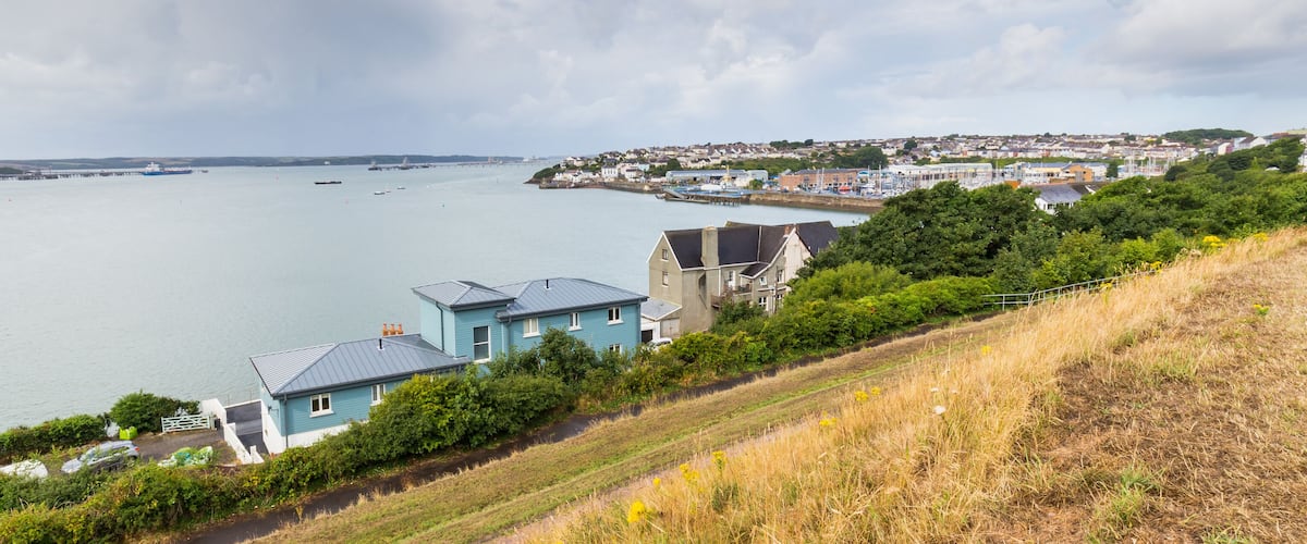 A coastal town with houses near a large body of water, a pier, and a bridge in the distance.