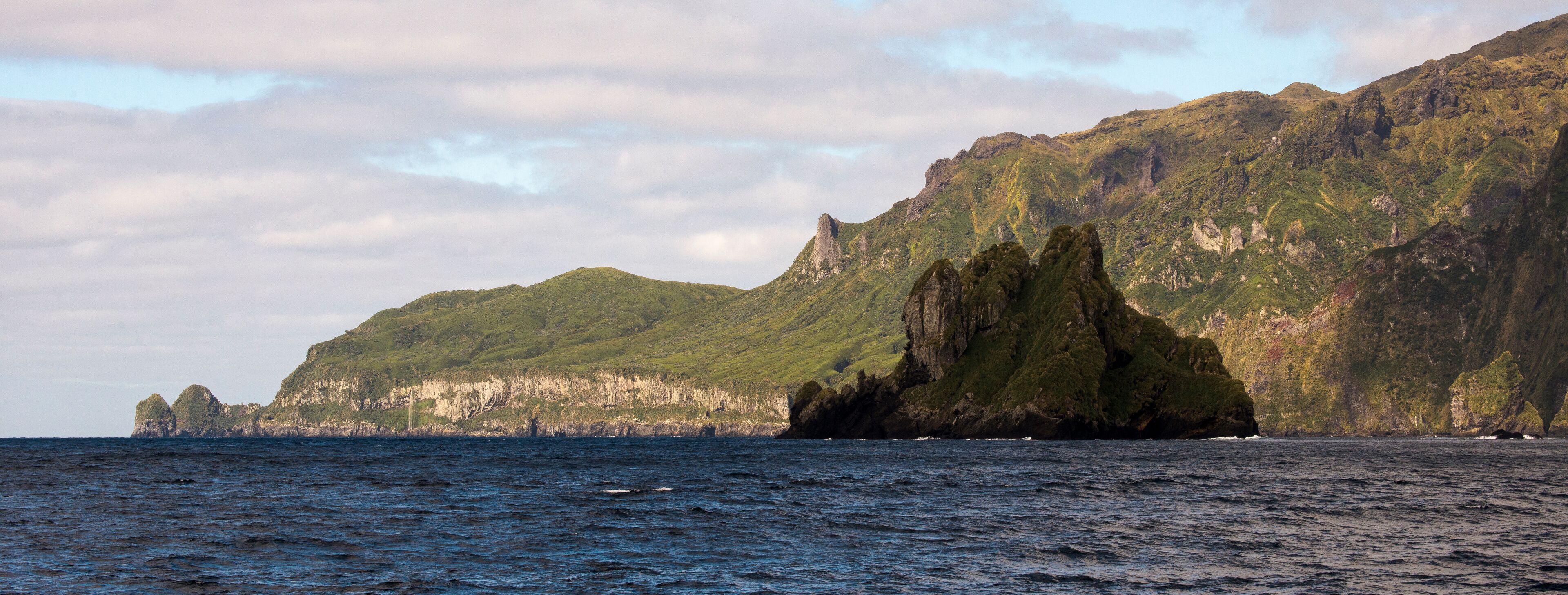 A rugged coastline with a prominent rock formation and lush green hills in the background.